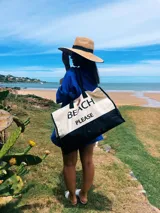 Bolso de playa grande de lona color crema con base negra y asas de soga negras. Estampado frontal con la frase "Beach Please" en letras negras.