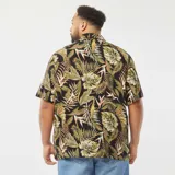 Camisa de manga corta con estampado de hojas tropicales en tonos verdes y marrones sobre fondo negro. Tiene cuello clásico, corte recto y cierre frontal con botones.