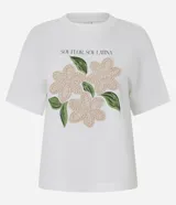 Remera blanca de algodón, con cuello redondo y manga corta. Presenta un estampado frontal con la frase "SOY FLOR. SOY LATINA" y un diseño floral con flores en relieve y hojas bordadas.
