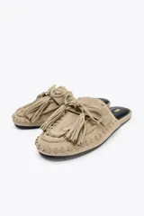 Zapato plano destalonado tipo mule, color beige, confeccionado en serraje. Presenta antifaz y lazo con borlas en la parte delantera.