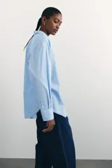 Camisa celeste de algodón suave, de corte holgado, con cuello clásico, botones frontales, canesú en la espalda, hombros caídos, bolsillo de parche en el pecho, mangas largas con puños abotonados y bajo redondeado.