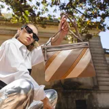 Sobre de mano con diseño de franjas diagonales en tonos beige y marrón, con cierre metálico frontal. Incluye correa desmontable para usar como bandolera.