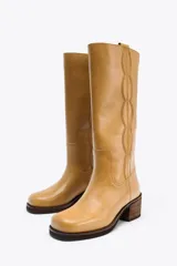 Bota alta de cuero color camel, con caña recta y punta redondeada. Presenta pespuntes decorativos verticales en la parte delantera y un tacón ancho de madera.