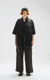 Camisa negra de manga corta, corte oversize, confeccionada en poplin 100% algodón. Presenta dos bolsillos delanteros, uno con solapa y otro con ojal y botón.