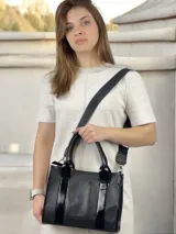 Bolso negro de cuero con detalles charolados, doble asa corta y correa larga de tela con el nombre de la marca.