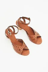 Sandalia de cuero color caramelo con taco bajo de madera. Presenta un diseño calado con bordes festoneados y perforaciones, y tira ajustable con hebilla en el tobillo.