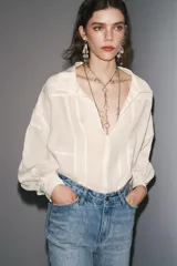 Camisa color crudo confeccionada en hilatura con mezcla de ramio, de corte holgado, con cuello solapa y escote pico. Tiene mangas largas con puños y detalle de tejido con pliegues.