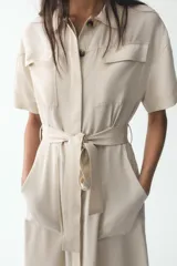 Vestido camisero largo color beige, de manga corta y cuello solapa. Tiene bolsillos de parche en el pecho, bolsillos laterales ocultos y cinturón de la misma tela.