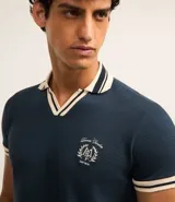 Remera polo comfort color crudo, de manga corta con ribete verde y cuello en V con ribete verde. Tiene un pequeño logo bordado en el pecho.