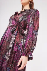 Vestido largo cruzado con estampado floral en tonos fucsia, negro, rojo y blanco. Tiene escote en V con volantes, mangas largas abullonadas y lazo para ajustar en la cintura.