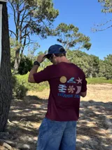 Remera de manga corta color bordó con un estampado gráfico en la espalda que incluye un sol amarillo, una ola azul, una estrella de mar blanca, un símbolo estilizado en azul y la frase "VITAMIN SEA" en azul.