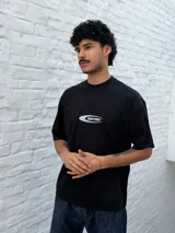 Remera negra de corte oversize con un pequeño gráfico ovalado en el pecho que contiene la palabra "OSTEN" en tipografía futurista.