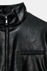 Cazadora corta negra de efecto piel, con cuello subido y manga larga. Tiene bolsillos delanteros con cremallera metálica y cierre frontal con cremallera.