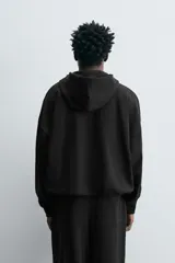 Sudadera negra con capucha, corte boxy, cierre frontal con cremallera, bolsillos laterales y puños elásticos.