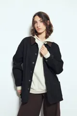 Sobrecamisa tipo barn jacket color negro, de algodón con elastano, con silueta relajada y estructura firme. Presenta cierre frontal con botones metálicos, cuello marrón, bolsillos aplicados y pespuntes visibles en color marrón.