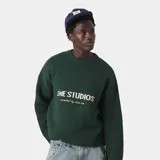 Sweater oversize color verde oscuro con cuello redondo y puños y dobladillo acanalados. Presenta el logo "EME STUDIOS" bordado en blanco en el pecho y la frase "curated by ben ice" debajo.