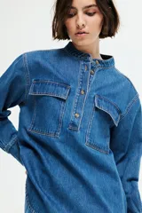 Camisa de denim azul de corte holgado, con cuello mao, tapeta de botones, hombros caídos, mangas largas con puños abotonados y dos bolsillos superiores con solapa.