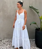 Vestido largo blanco de algodón con tirantes finos, escote en V con cordones y borlas, y detalles de encaje en el busto y en los volados de la falda.