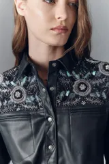 Camisa de símil cuero negra, de calce entallado, con cuello camisero y botones a presión con logo grabado. Presenta bordados florales con lentejuelas en los hombros y sobre los bolsillos frontales.