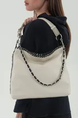 Cartera tipo shopper color arena marfil, de cuero vacuno graneado, con detalles en cuero negro brillante y apliques metálicos. Tiene doble manija regulable, correa ancha desmontable y reversible, y detalles de cadena entrelazada en cuero.