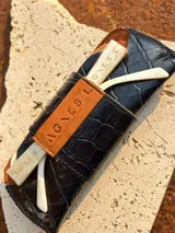 Estuche para lentes con textura símil cocodrilo color azul y cierre con tira de cuero marrón con el logo de la marca.