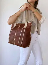 Bolso tipo tote de cuero marrón con costuras blancas a la vista y tiras decorativas. Asas de cuero color crema con detalle de trenzado. Forrada en tela.