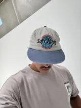 Gorro tipo gorra de béisbol, color gris claro con visera azul. Presenta un bordado frontal con el texto "Soft Rock Cafe" y un diseño de un globo terráqueo con detalles en rojo, azul y verde.