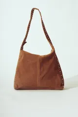 Bolso de hombro tipo hobo, color camel, confeccionado en gamuza o material similar con acabado afelpado. Presenta detalles de tachas metálicas plateadas a lo largo de los bordes laterales y en la correa de hombro.