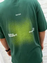 Remera oversize color verde oscuro con estampa minimalista en blanco que dice "living forward in motion" y un detalle gráfico punteado en verde lima en el pecho.