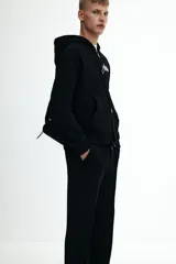 Pantalón tipo jogger negro, de corte recto y silueta clásica, confeccionado en tejido de punto de mezcla de algodón. Presenta pliegue cosido frontal, cintura elástica con cordón ajustable y bolsillos laterales y trasero insertado.