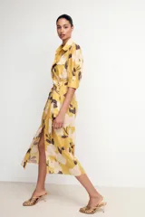 Vestido midi camisero color amarillo con estampado de flores blancas y hojas marrones. Tiene cuello solapa, mangas 3/4 con puños, cinturón de lazo ajustable y cierre frontal con botones.