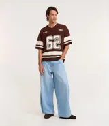 Remera oversized deportiva color marrón con cuello en V, rayas blancas en las mangas y en la parte inferior, y el número "62" estampado en blanco en el frente. Incluye el bordado "Humanity" y un logo de globo terráqueo.