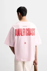 Camiseta rosa de corte holgado, cuello redondo y manga corta. Estampado frontal con texto en rojo "Amalfi Coast" y estrellas.