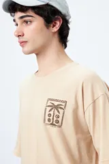 Remera color beige de corte amplio con mangas cortas y estampado de una postal con una palmera en el pecho.