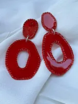 Aros colgantes con forma geométrica irregular, color rojo. El aro superior es pequeño y redondeado, mientras que el aro inferior es grande y con un agujero en el centro.