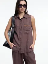 Camisa sin mangas color marrón confeccionada en lino, con cartera abotonada en la parte baja de la espalda, bolsillo delantero y bordado tipo pespunte en color contraste tanto en el frente como en el canesú de la espalda.