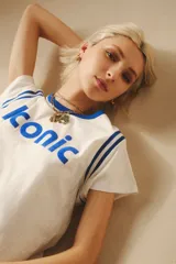 Remera blanca de algodón con cuello redondo y mangas cortas con ribetes azules. Estampado frontal con la palabra "Iconic" en color azul.