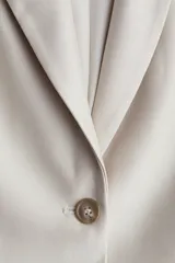 Blazer color beige de corte entallado, con solapas de pico, cierre de un botón, hombreras, bolsillos delanteros de parche, puños con abertura y botón, y abertura trasera.