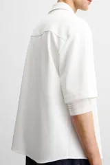Sobrecamisa color beige de tejido elástico, con cuello solapa, manga corta y bolsillos de parche en el pecho. Cierre frontal con botones a presión.