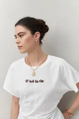 Remera blanca de manga corta con bordado frontal "C’est la vie", calce relajado y frunces laterales.