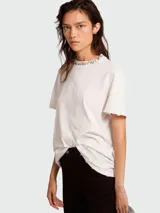 Remera de algodón color blanco roto, de corte holgado y manga corta con bordes deshilachados. Presenta un cuello redondo adornado con cristales cabujón engarzados.