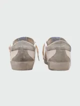 Championes Golden Goose modelo Super Star, color blanco con detalles en gris y suela color crema. Presentan efecto desgastado y estrella distintiva de la marca en el lateral.