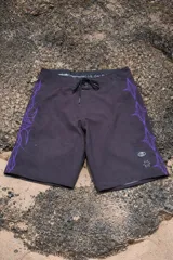 Short de baño negro con cordón de ajuste blanco en la cintura. Presenta un estampado vertical de figuras abstractas en color violeta y un logo estilizado en blanco en el lateral derecho. En el ruedo inferior izquierdo tiene un pequeño logo circular y una estrella de cinco puntas.