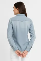 Camisa de denim celeste, corte entallado, con cuello clásico, cierre frontal con botones a presión, mangas largas con puños abotonados, dos bolsillos con solapa abotonada en el pecho y canesú en la espalda.