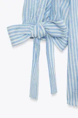 Camisa celeste y blanca a rayas verticales, confeccionada con lino e hilo metalizado. Tiene cuello solapa, mangas 3/4 con lazos en los puños y cierre frontal con botones ocultos por solapa.
