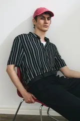 Camisa de manga corta con rayas verticales blancas sobre fondo negro, cuello inglés, tapeta con botones y bolsillo de parche en el pecho. Corte recto y holgado.