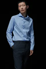 Camisa celeste de algodón con cuello italiano, tapeta clásica y canesú en la espalda. Mangas largas con tapeta y botón, y cierre ajustable de botón en los puños. Bajo levemente redondeado. Corte ajustado que realza el contorno del cuerpo creando una silueta entallada.