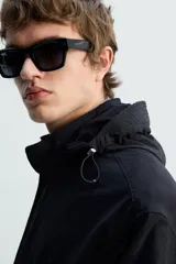 Parka negra de tejido técnico ligero, corte regular, cuello alto con capucha ajustable, manga larga con puños ajustables, cintura ajustable con cordón interno, bolsillos de solapa en la cadera, abertura en la espalda y cierre frontal con cremallera oculta y botones a presión.