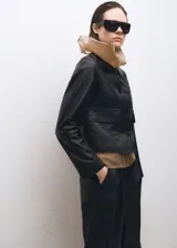 Chaqueta negra de cuero ovino con cuello de solapa, diseño recto, dos bolsillos de parche con solapa en la parte delantera y cierre delantero con botones.