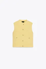 Chaleco amarillo de punto con cuello redondo y sisas amplias. Tiene cierre frontal con botones dorados y dos bolsillos de parche con botón.